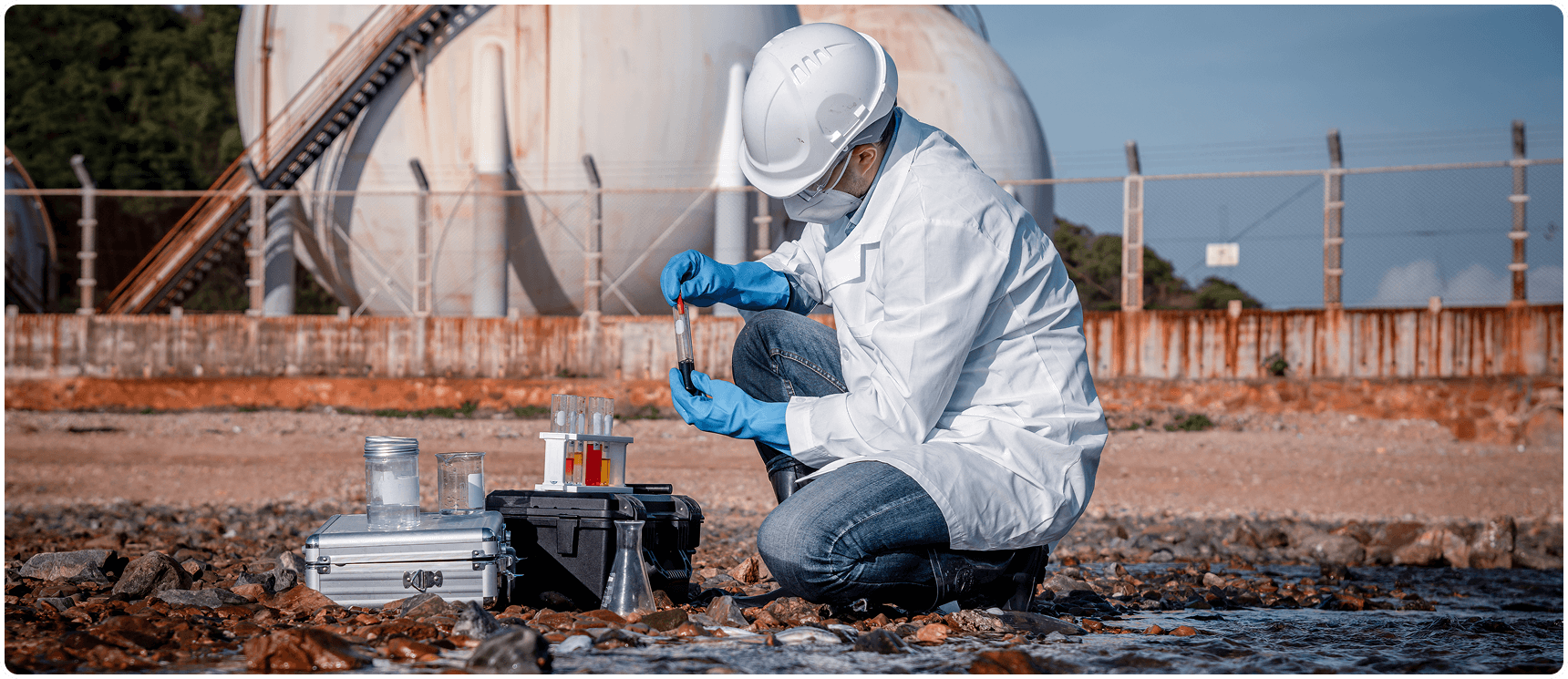 Scientist collecting water sample near industrial site.